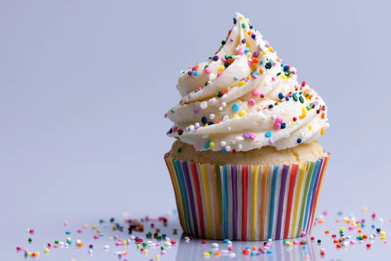 Single cupcake with white frosting and colorful sprinkles in rainbow-striped paper liner on reflective surface