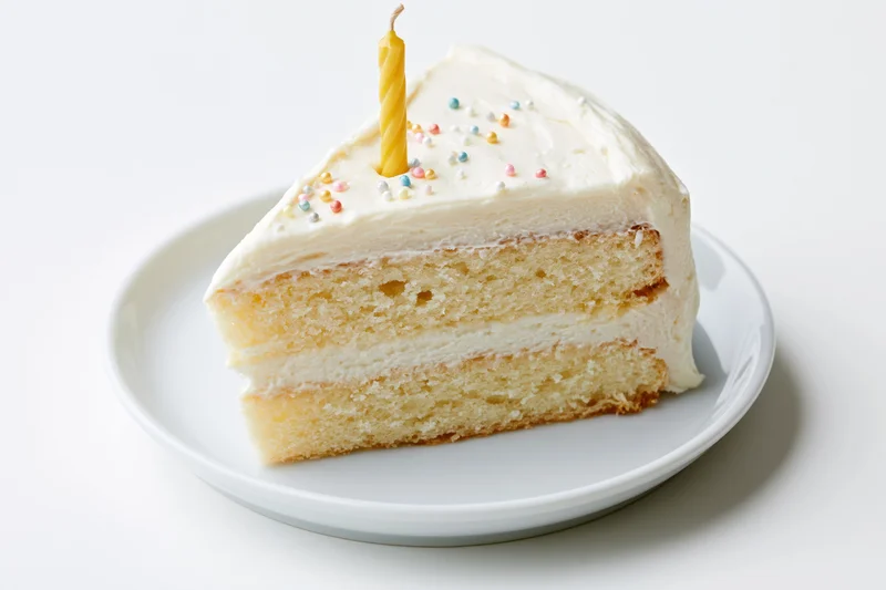 Slice of white-frosted layer birthday cake with yellow candle and sprinkles on white plate