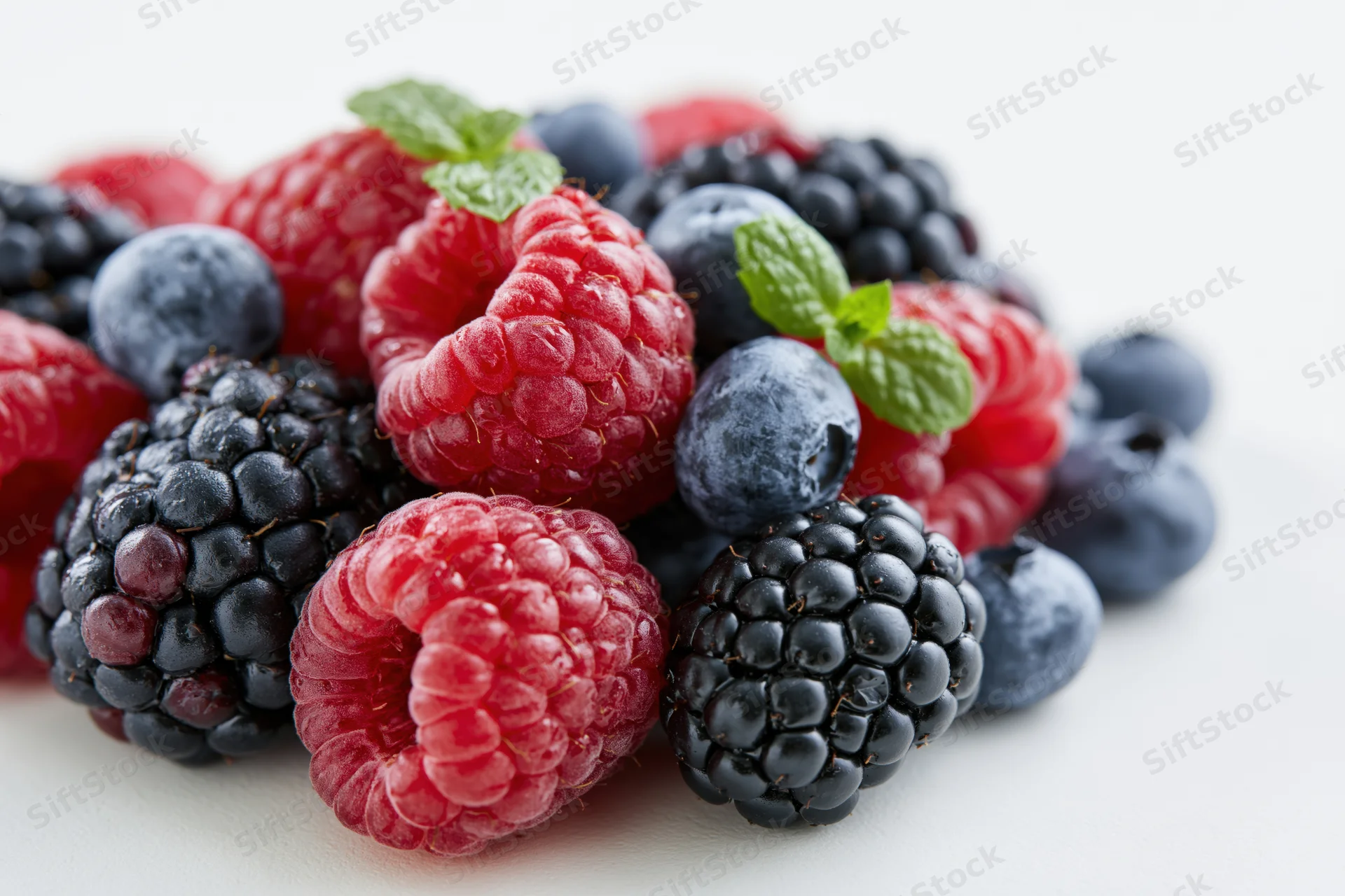 A pile of fresh raspberries, blackberries and blueberries with mint leaves on a white surface