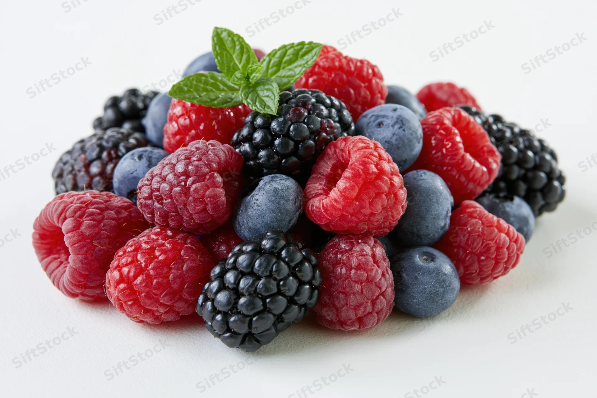 A pile of fresh raspberries, blackberries and blueberries with a mint sprig on a white surface
