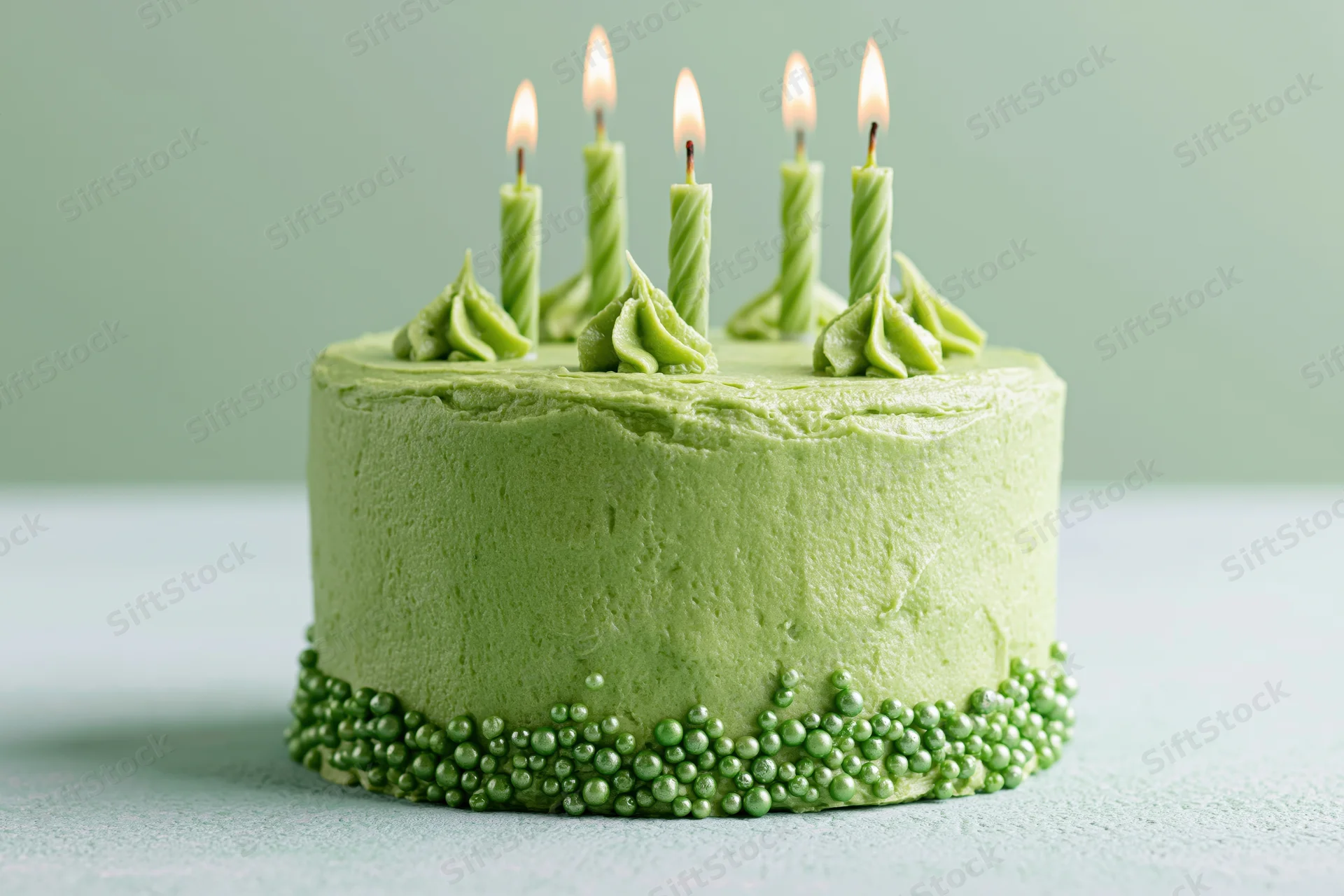 Small round green frosted birthday cake with lit candles and sprinkles on a light surface