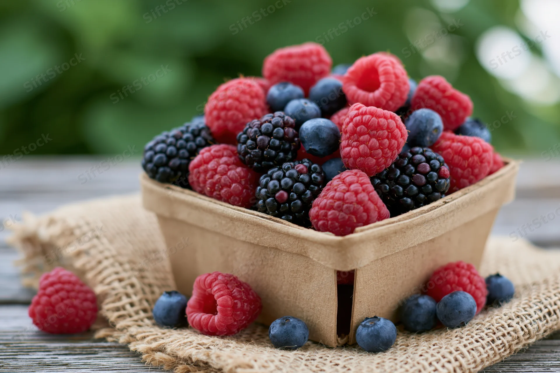A cardboard basket overflowing with raspberries, blueberries and blackberries on a burlap cloth over a wooden table outdoors