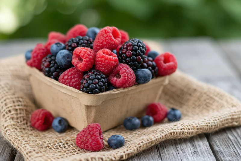 Mixed berries overflowing from a cardboard basket on burlap over wooden table