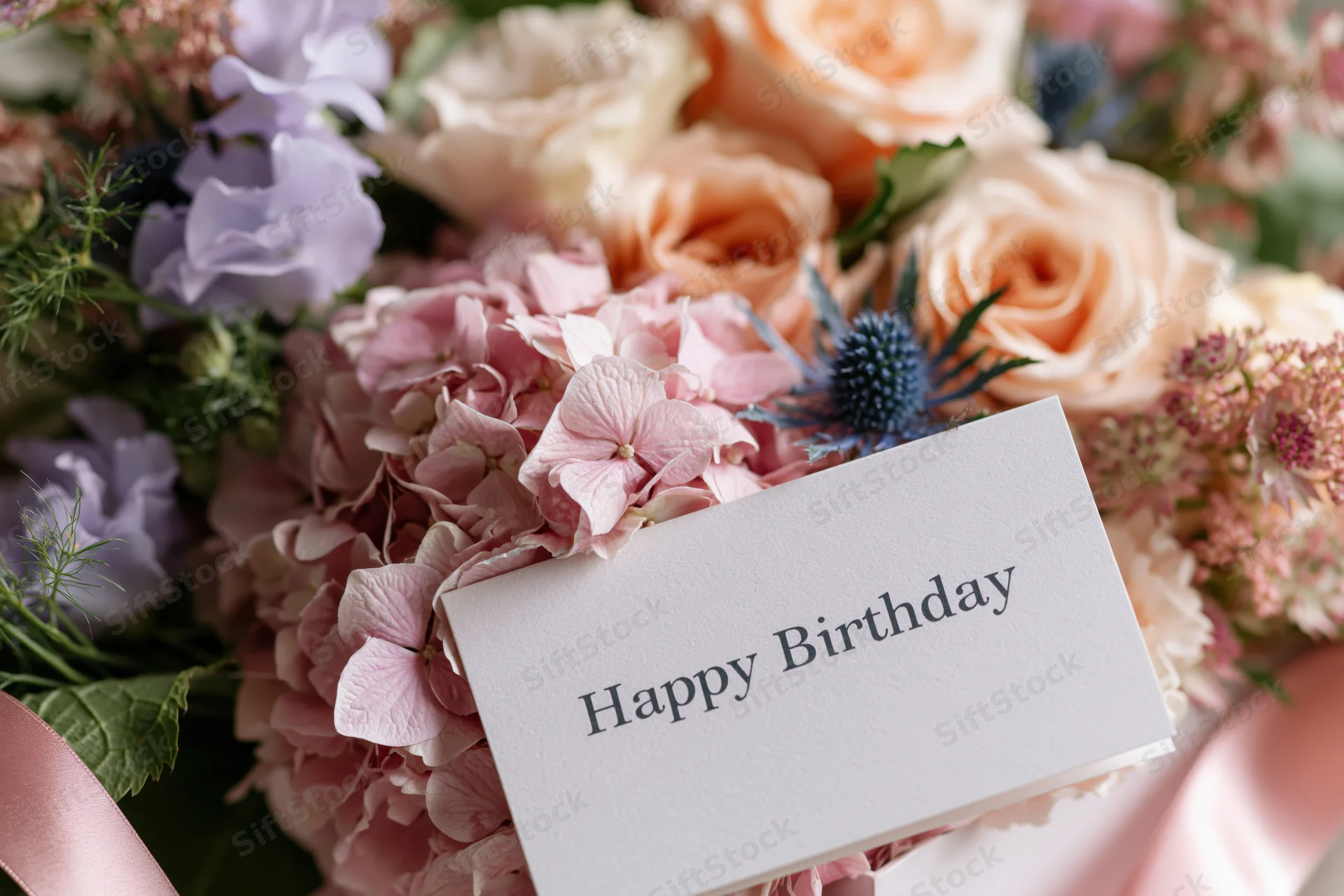 Birthday card with happy birthday text nestled in a bouquet of pink and peach roses, hydrangeas, and blue thistles, tied with a pink ribbon