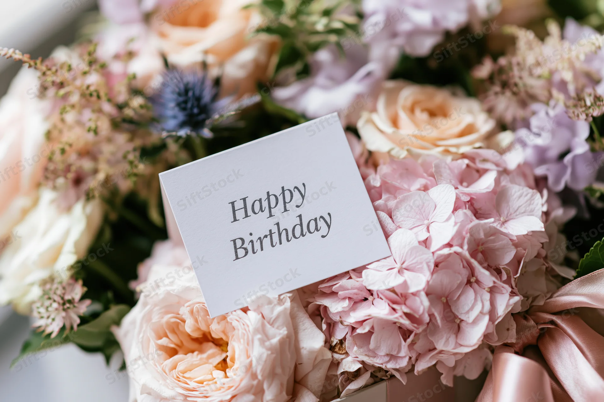 Birthday card with happy birthday text nestled in a bouquet of pink and peach roses and hydrangeas with ribbon