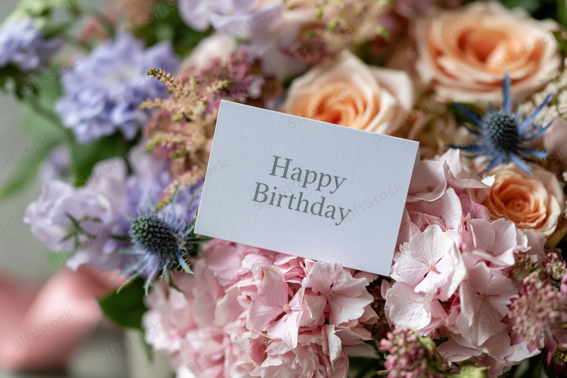 Birthday card with happy birthday text nestled in a bouquet of pink roses, hydrangeas, and blue thistles