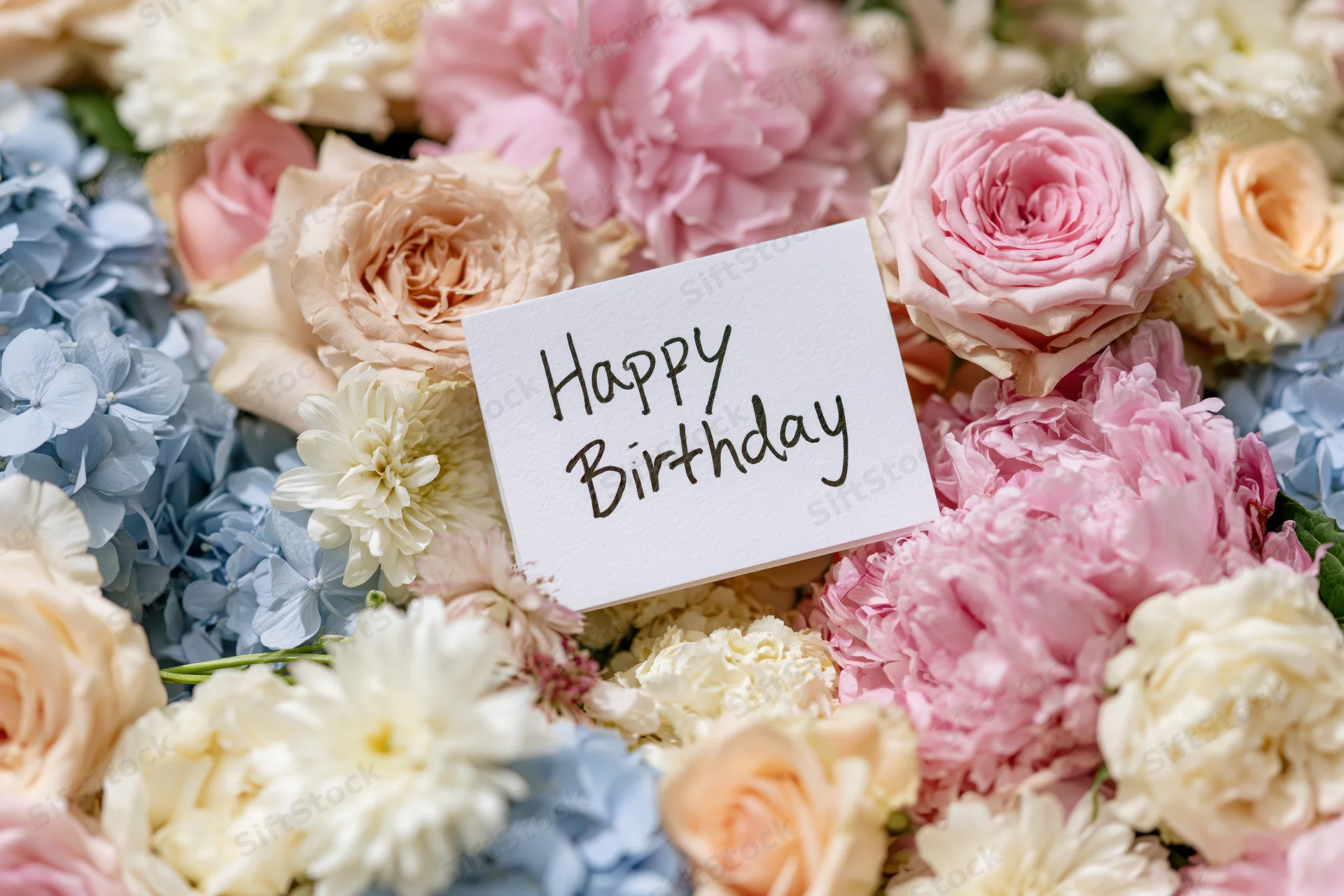A white card with handwritten happy birthday message placed among pink roses, blue hydrangeas, and cream peonies in a floral arrangement