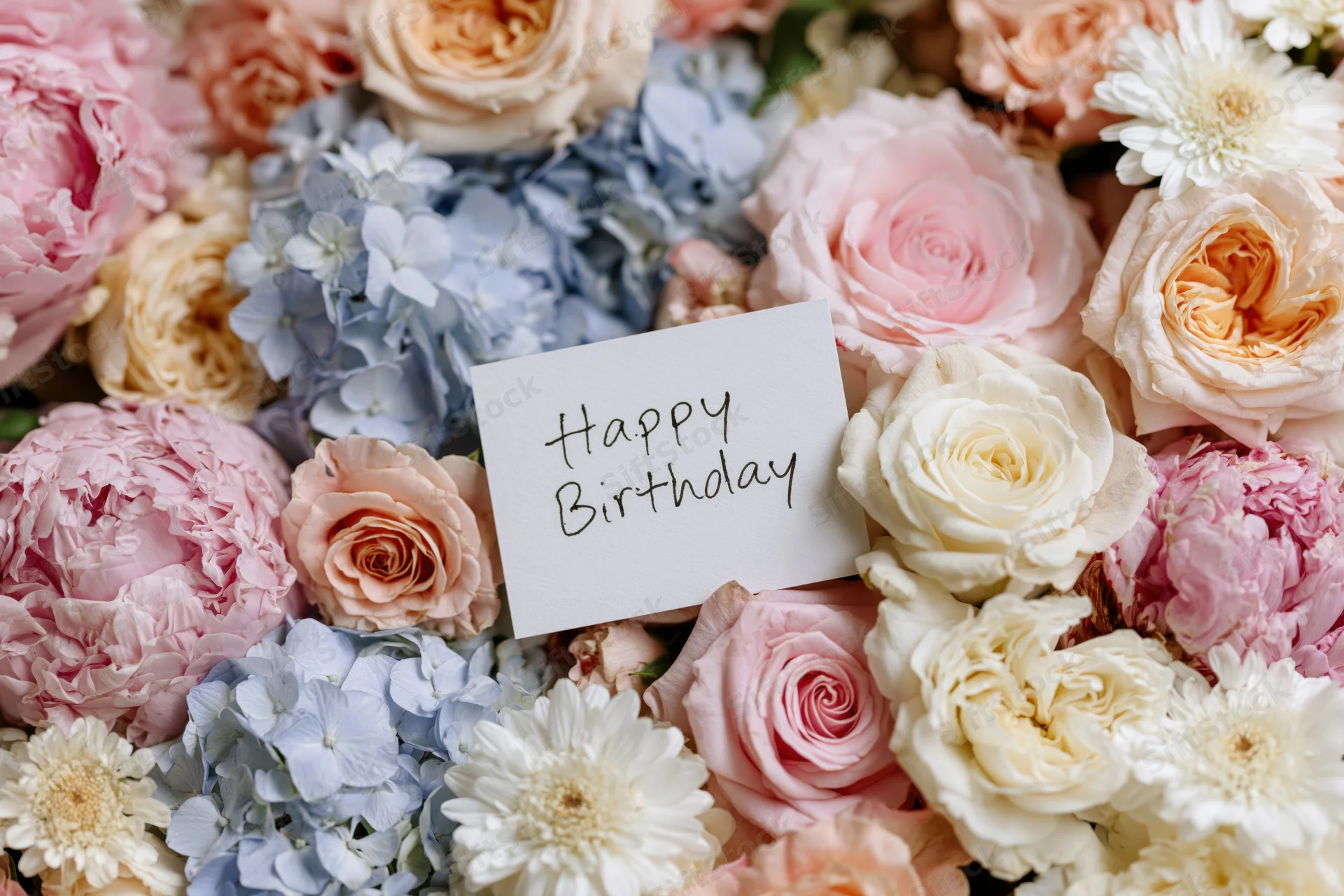 A bouquet of pink, peach, cream and blue flowers with a white card reading happy birthday nestled among the blooms