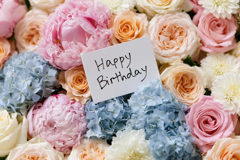 A bouquet of pink, peach, cream and blue flowers with a white card reading happy birthday nestled among the blooms