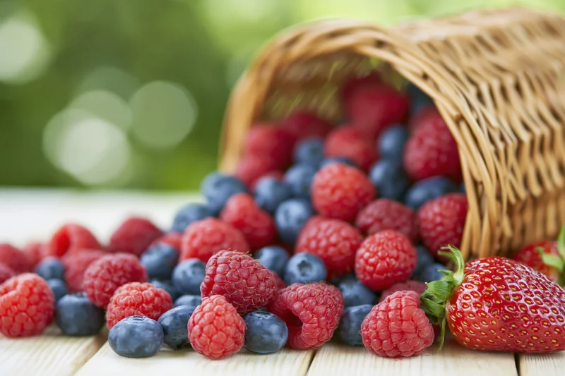 Spilled raspberries, blueberries and strawberries from a tipped wicker basket on a wooden table