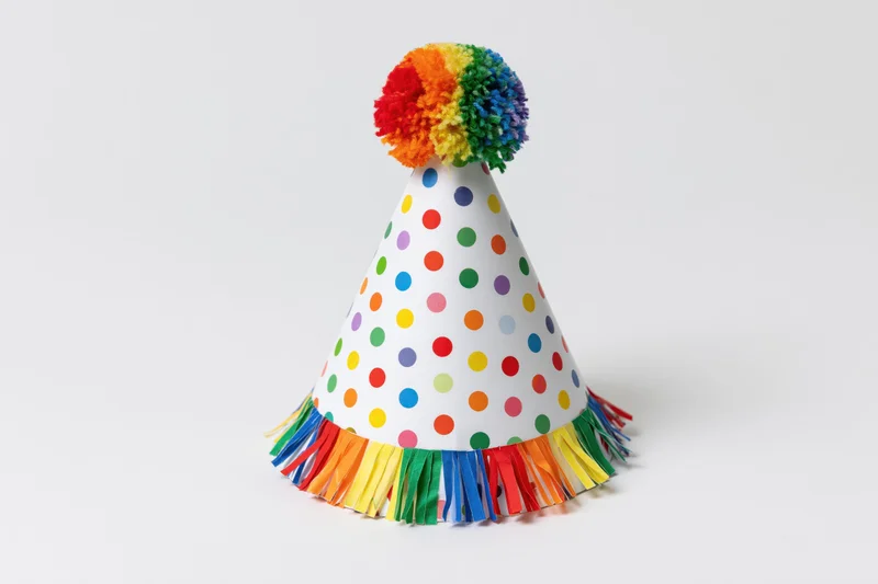White conical party hat with rainbow pompon and colorful fringe on plain white background