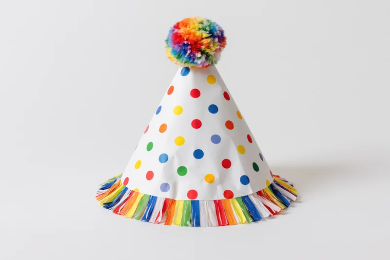 White conical party hat with colorful polka dots and rainbow fringe on plain background