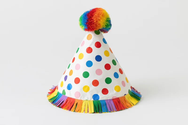 White conical party hat with rainbow pompon and colorful fringe on plain background