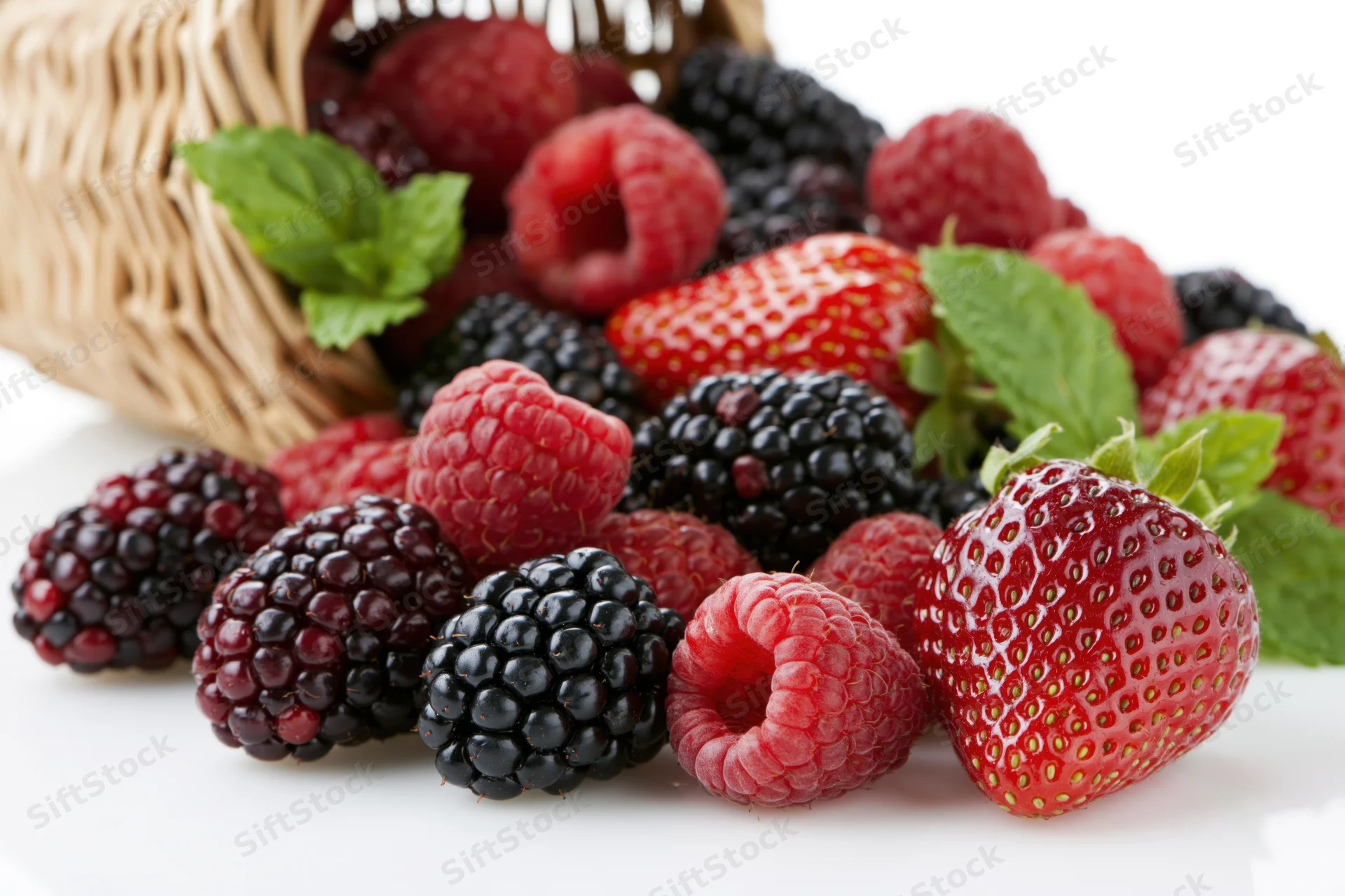 Wicker basket tipped over spilling fresh strawberries raspberries and blackberries onto white surface