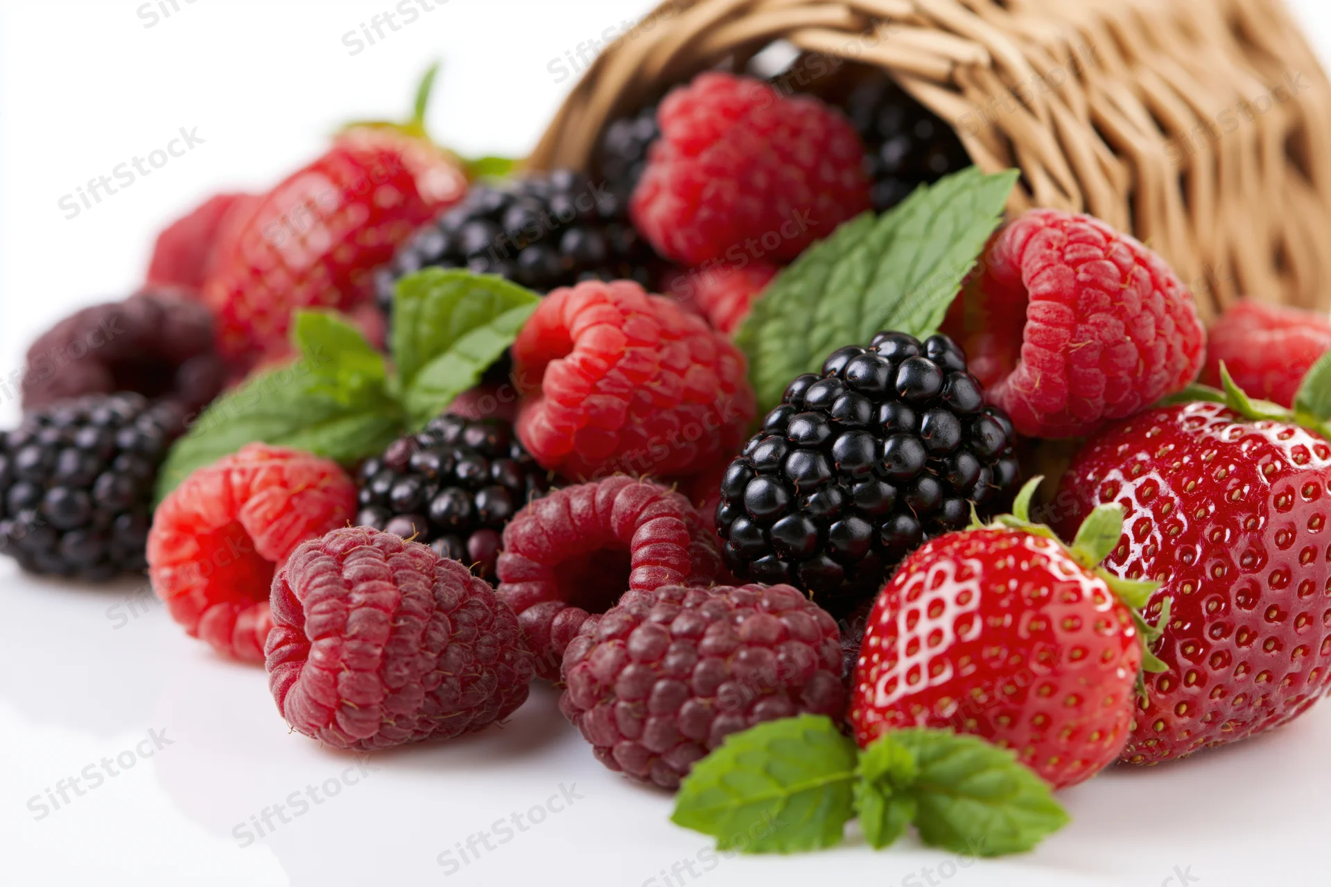 Ripe raspberries, blackberries and strawberries spilling from a wicker basket onto a white surface with mint leaves scattered around