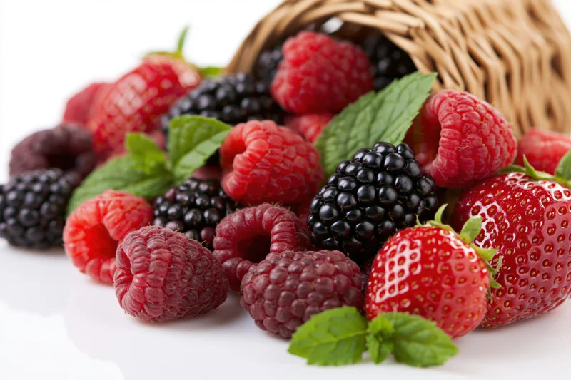 Ripe raspberries, blackberries and strawberries spilling from a wicker basket onto a white surface with mint leaves scattered around