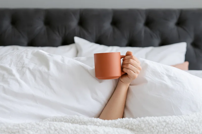 Woman's arm reaching from under white bedding holding a terracotta mug in bed