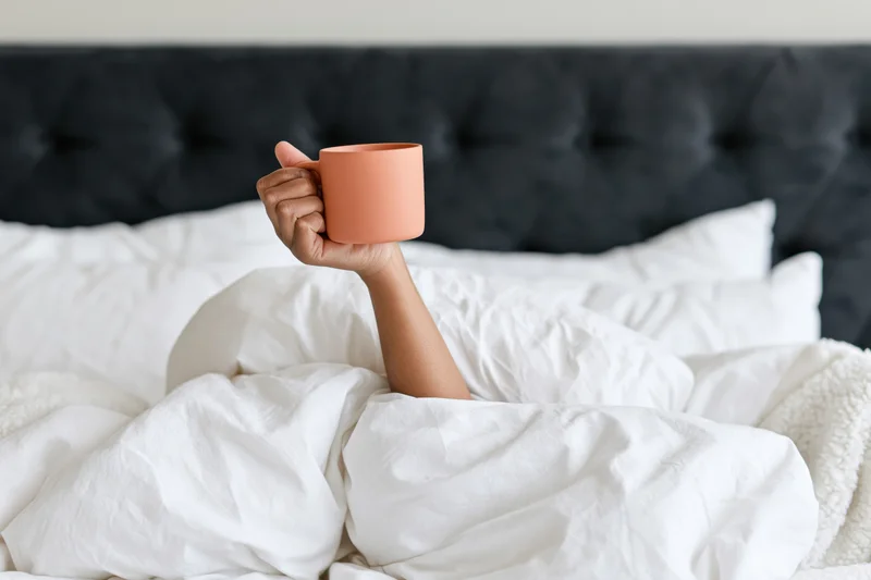Woman’s arm reaching from under white bedding holding a peach mug in bed