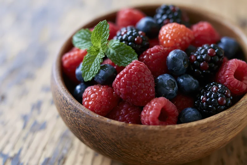 Wooden bowl filled with fresh raspberries blueberries blackberries and mint on a rustic wooden surface