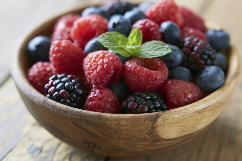 Wooden bowl filled with fresh raspberries blueberries and blackberries garnished with mint on a rustic table