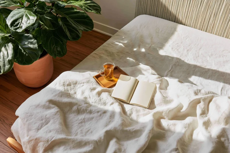 Unmade bed with rumpled white linen sheets, open notebook and tea cup on wooden tray, beside potted fiddleleaf plant in sunlit bedroom