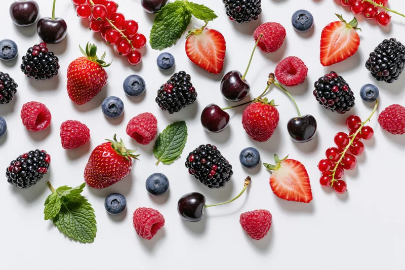 Assorted fresh berries and cherries scattered on white surface with mint leaves