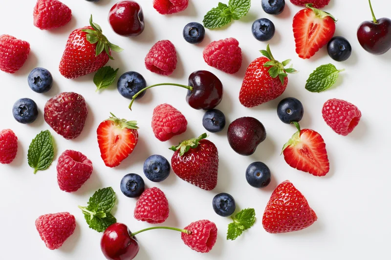Assorted fresh berries and cherries scattered on a white surface with mint leaves