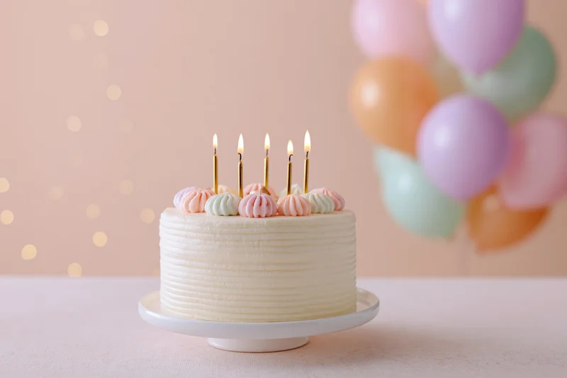 Birthday cake with lit candles on a white cake stand, pastel balloons in background
