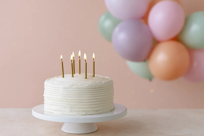 White frosted birthday cake with lit candles on a stand beside pastel balloons on a table