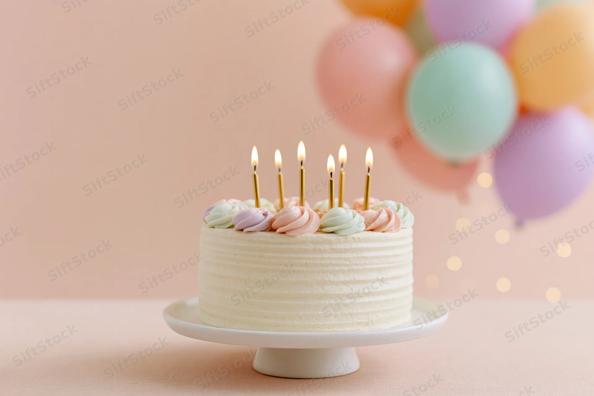 Birthday cake with lit candles on a white cake stand, pastel balloons in background