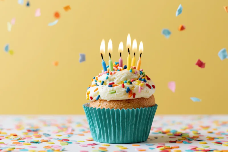 Birthday cupcake with lit multicolored candles and sprinkles on a table with falling confetti