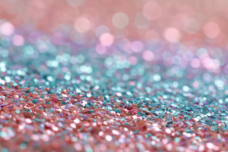 Close-up of scattered rainbow glitter and sequins on a soft pastel surface with bokeh background