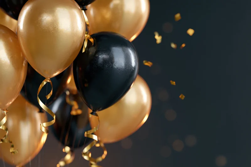 Cluster of gold and black metallic balloons with ribbons and confetti floating against dark background