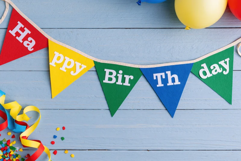 Colorful happy birthday pennant banner strung across a blue wooden surface with balloons, streamers and confetti