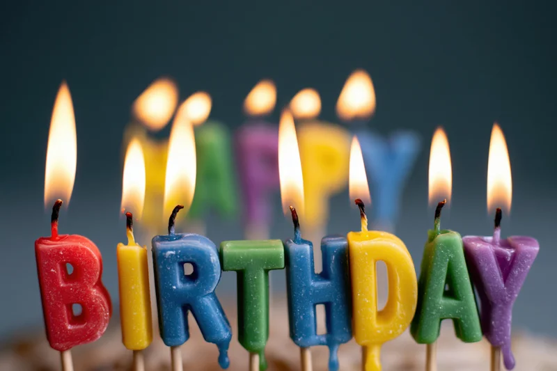 Colorful letter-shaped birthday candles spelling birthday on a cake, lit with flames, close-up view against a dark background with shallow depth of field and copyspace on the sides