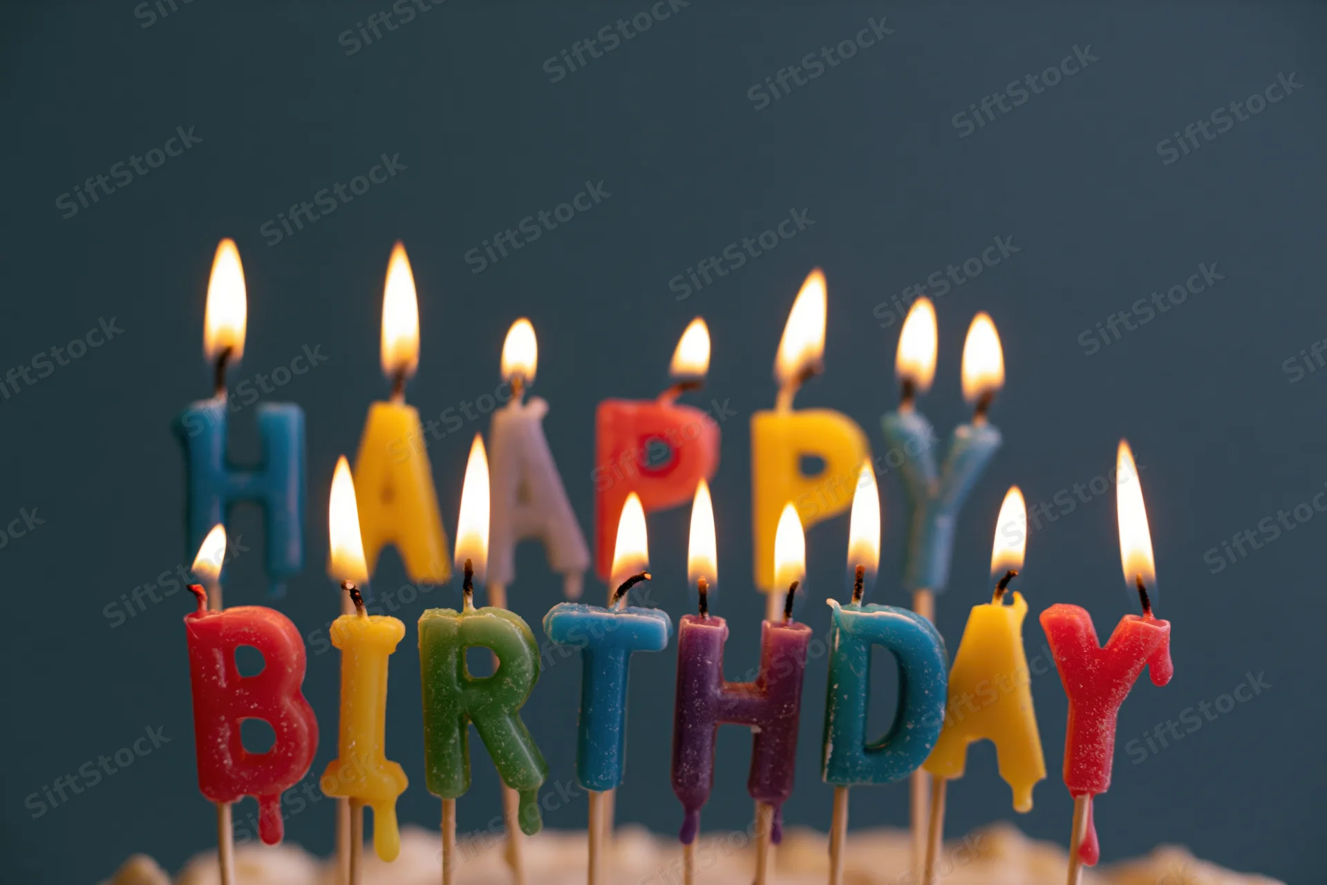 Colorful letter-shaped birthday candles spelling happy birthday on a cake with lit flames against a dark background