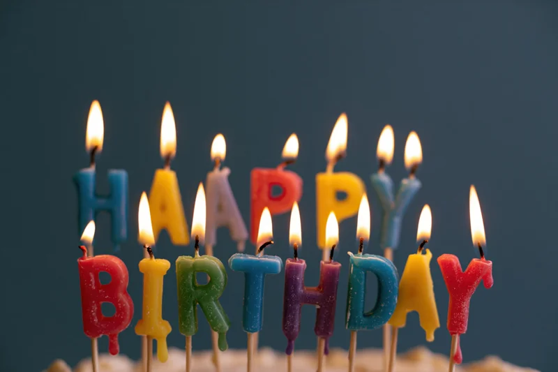 Colorful letter-shaped birthday candles spelling happy birthday on a cake with lit flames against a dark background