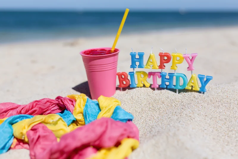 Colorful letter candles spelling happy birthday stuck in sand on beach with pink cup and straw nearby