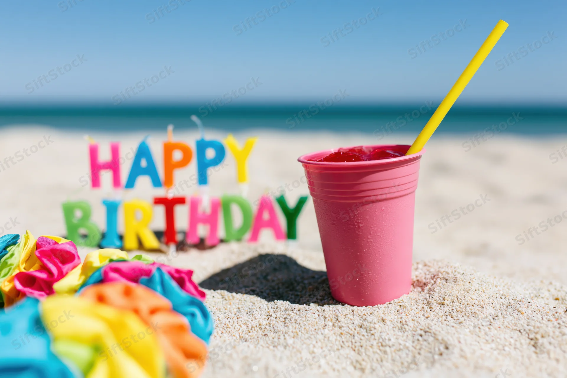 Colorful letter candles spelling happy birthday in sand on beach with pink cup and straw nearby