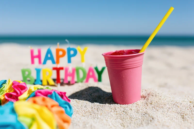 Colorful letter candles spelling happy birthday in sand on beach with pink cup and straw nearby