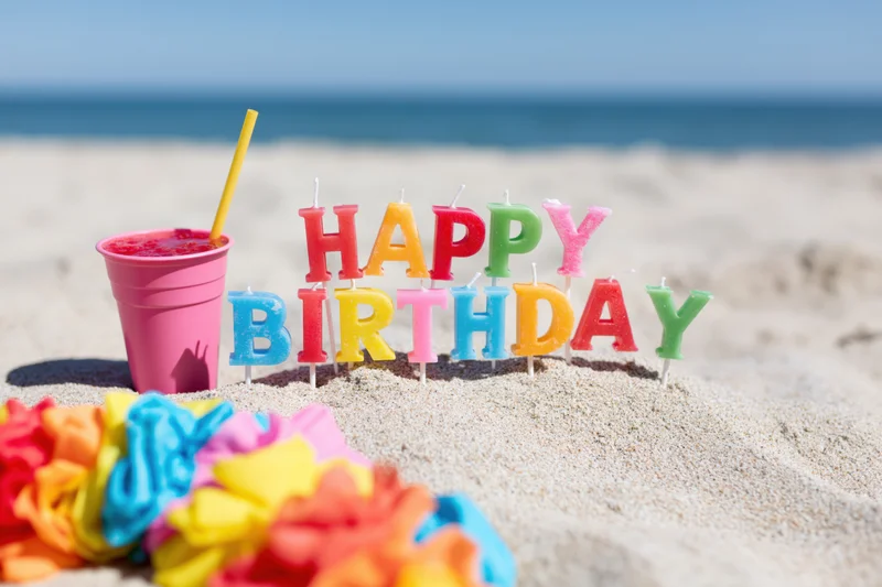 Colorful letter candles spelling happy birthday stuck in sand on beach with drink and lei nearby