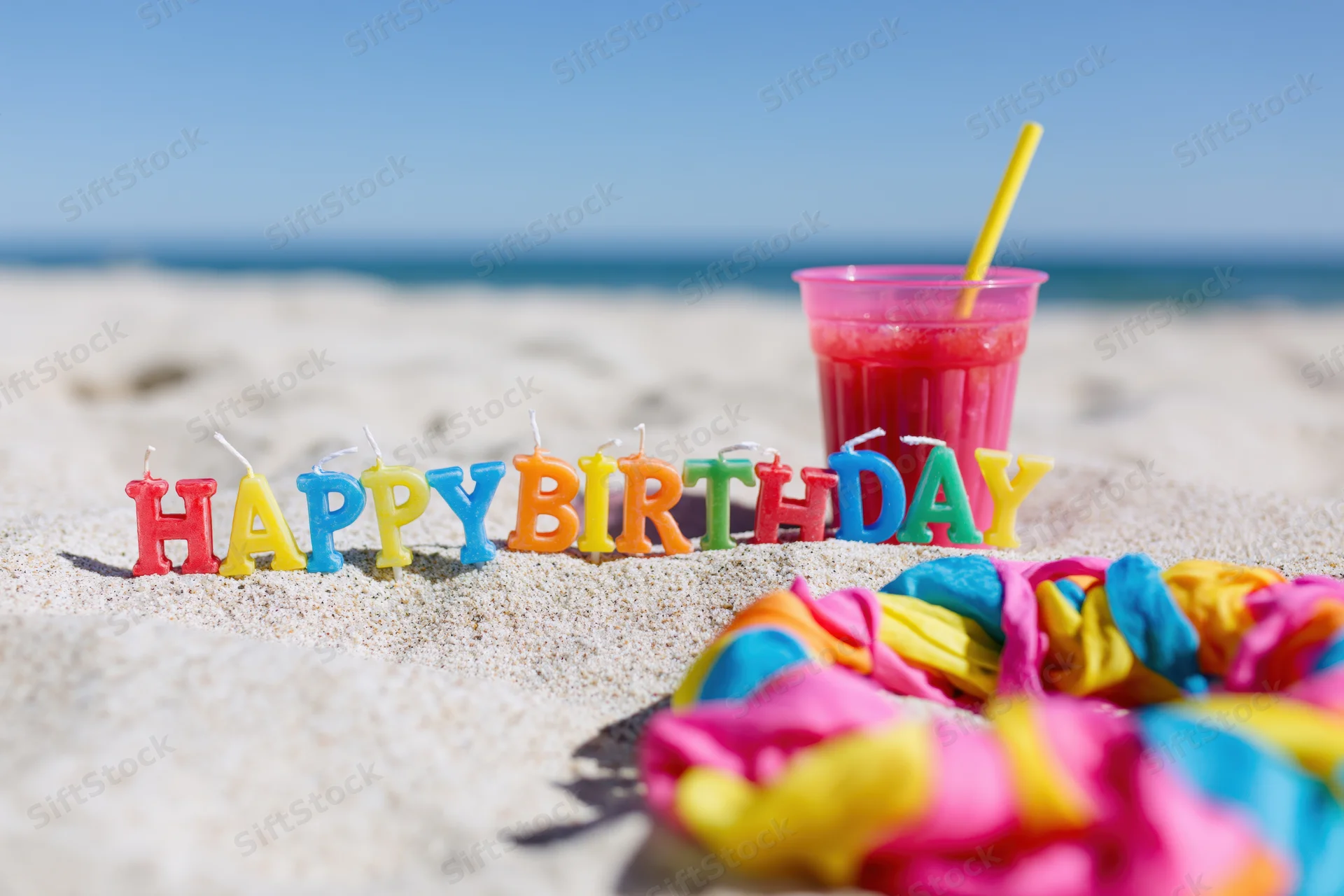 Colorful letter candles spelling happy birthday in sand on beach with drink and lei