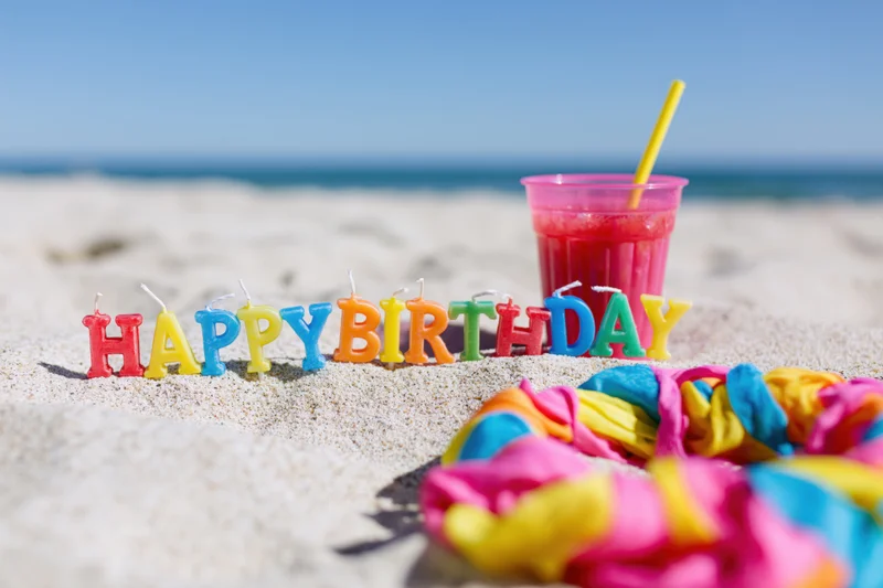 Colorful letter candles spelling happy birthday in sand on beach with drink and lei