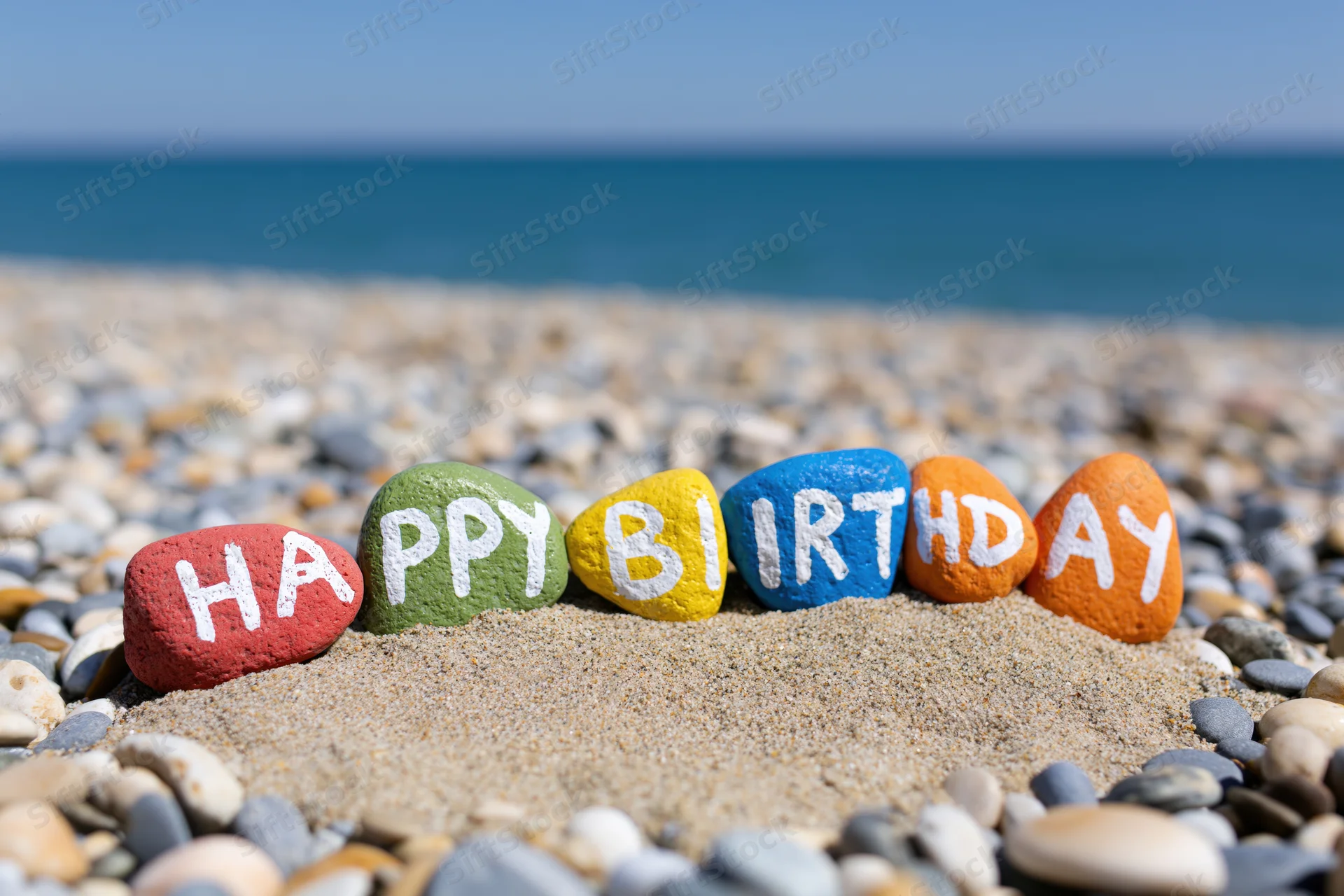 Colorful painted stones spelling happy birthday on sandy beach with ocean in background