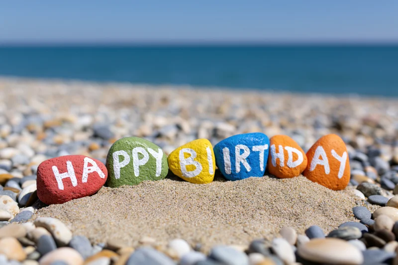 Colorful painted stones spelling happy birthday on sandy beach with ocean in background