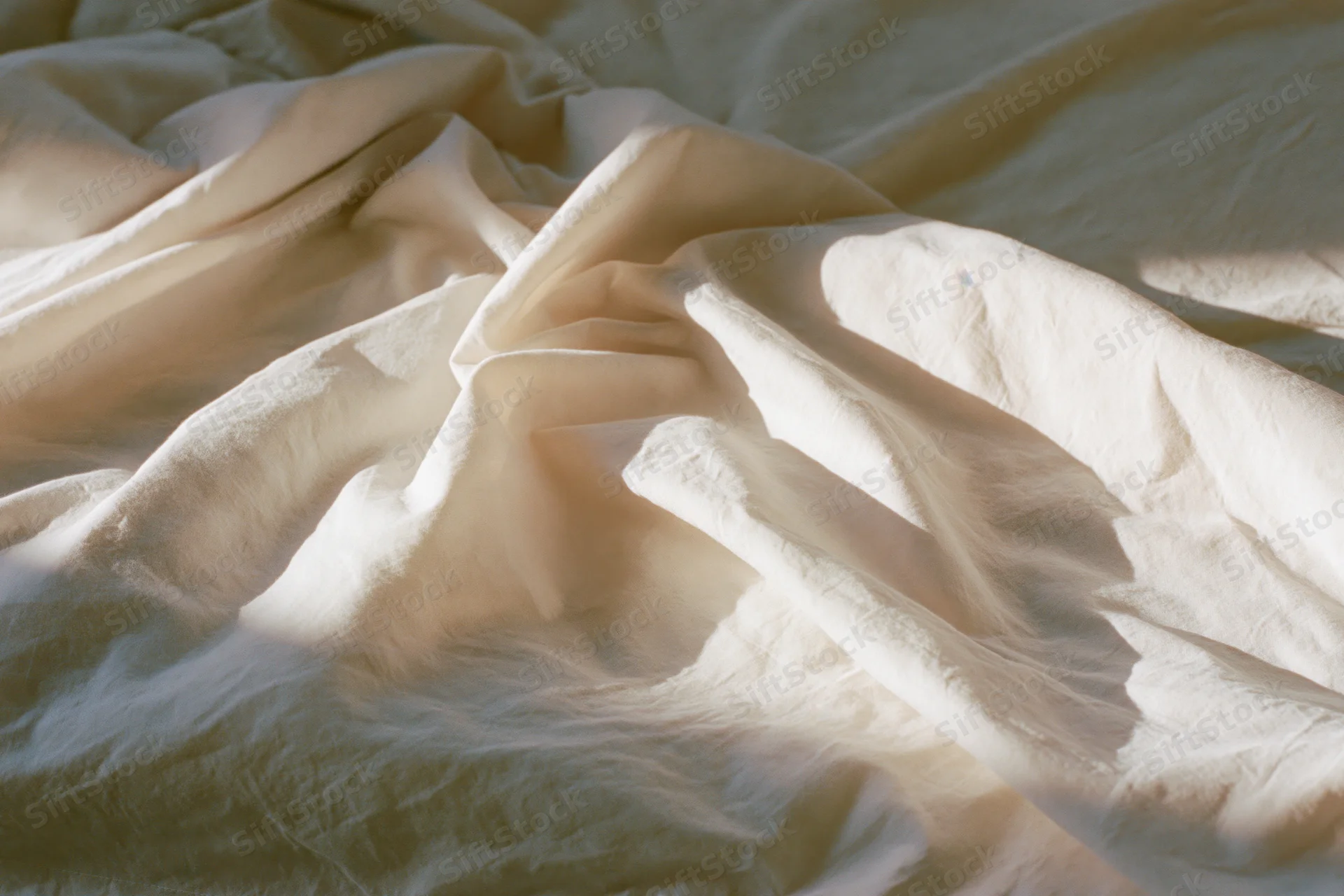 Crumpled white bedsheet draped over edge of unmade bed in soft morning light