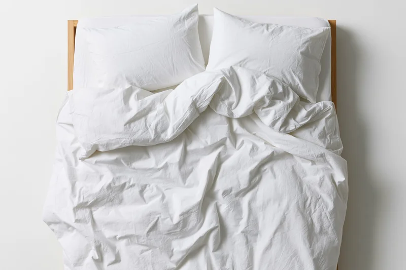 Unmade double bed with crumpled white cotton duvet and two pillows against white wall