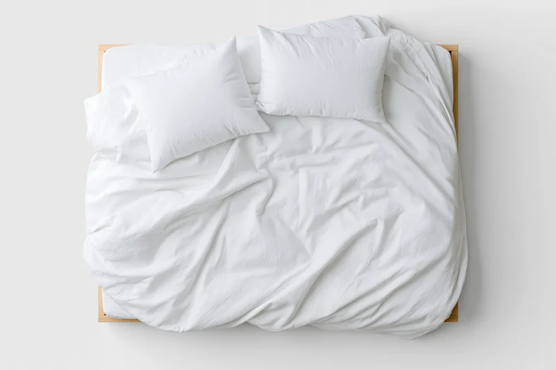 Overhead view of a double bed with rumpled white cotton duvet and two pillows on a wooden frame against a plain white wall