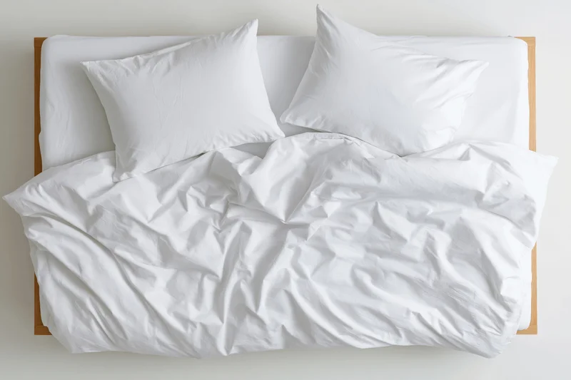 Overhead view of unmade double bed with white cotton duvet and two pillows on wooden frame