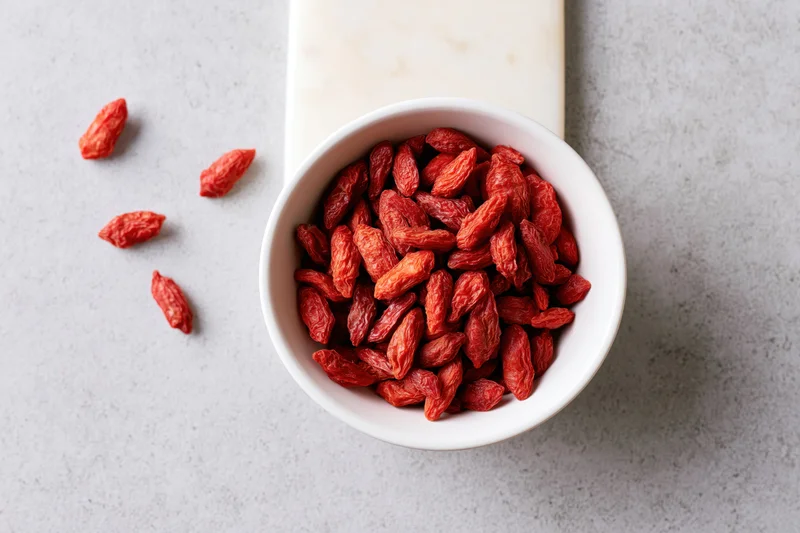 Dried goji berries in a white ceramic bowl on a gray countertop with a few scattered beside it